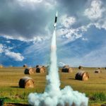 Small model rocket flying up in smoke after launch on green field with hay rolls