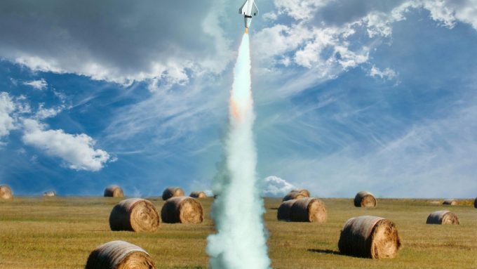 Small model rocket flying up in smoke after launch on green field with hay rolls