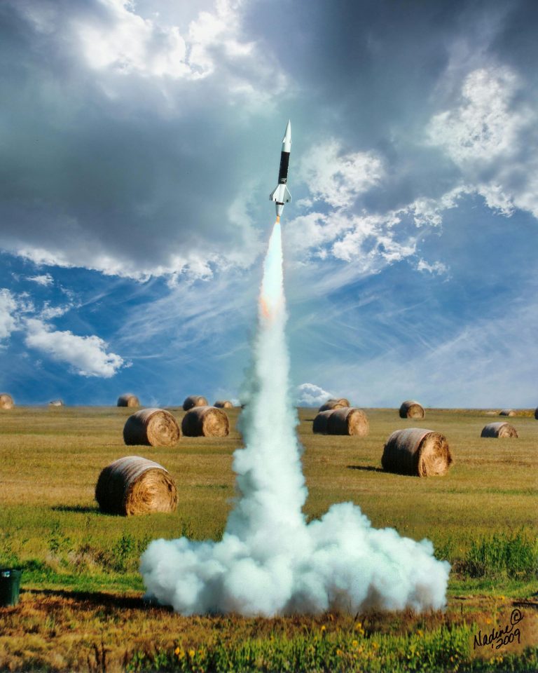 Small model rocket flying up in smoke after launch on green field with hay rolls